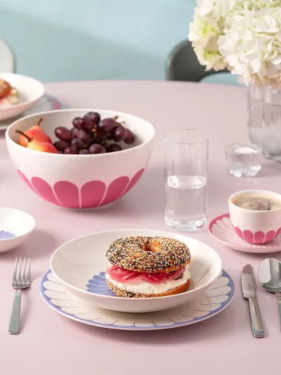 Ein runder rosa Tisch mit Tellern voller Essen, darunter ein belegter Bagel, eine Obstschale, Tassen Kaffee und Besteck. Ein runder rosa Tisch mit Tellern voller Essen, darunter ein belegter Bagel, eine Obstschale, Tassen Kaffee und Besteck. Im Hintergrund steht eine Vase mit weißen Blumen.