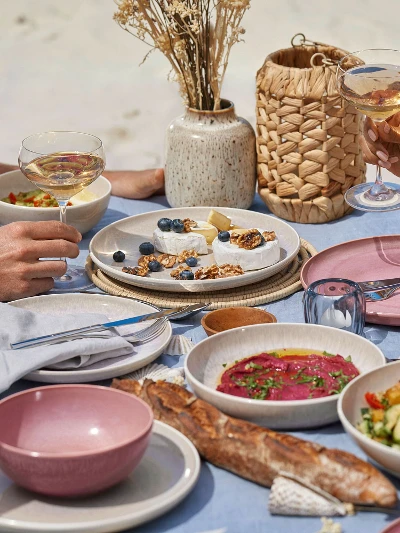 Een stel zit aan een tafel met borden van de Perlemor collectie van like. by Villeroy & Boch op het strand.