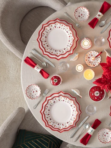 Festively decorated Christmas table with Toy's Delight tableware Top-down view of a round dining table elegantly set for Christmas with Villeroy & Boch's Toy's Delight porcelain plates, red napkins, candles, and a red poinsettia centerpiece next to a lit Christmas tree.