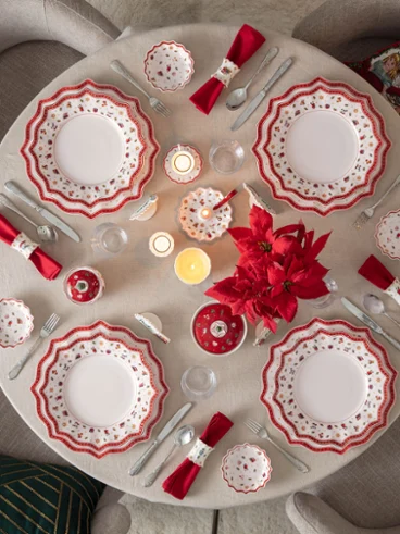 Festively decorated Christmas table with Toy's Delight tableware Top-down view of a round dining table elegantly set for Christmas with Villeroy & Boch's Toy's Delight porcelain plates, red napkins, candles, and a red poinsettia centerpiece next to a lit Christmas tree.