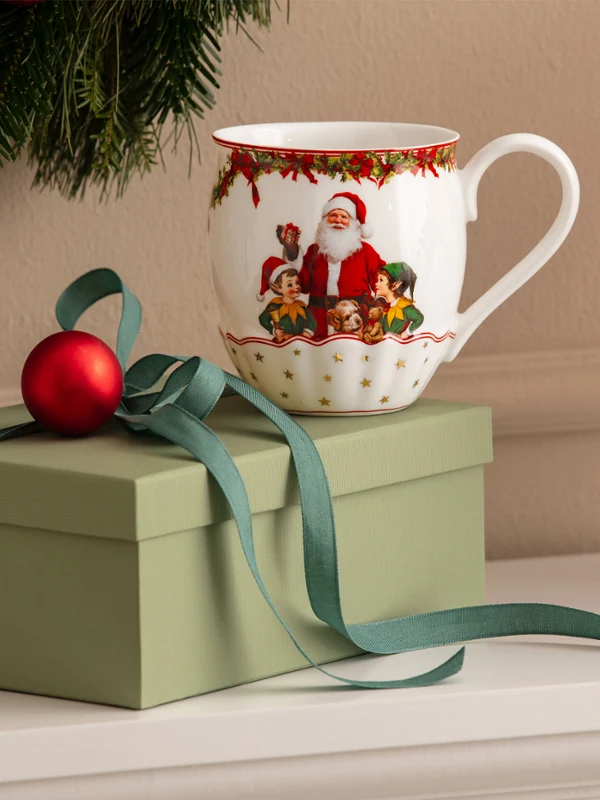 Annual Chistmas edition 2026 mug Eine grüne Geschenkbox mit einer Schleife, einem roten Christbaumschmuck und einer Tasse mit Weihnachtsmann-Motiv von Villeroy & Boch vor einem weißen Hintergrund, im Hintergrund sind weihnachtliche Dekorationen zu sehen.