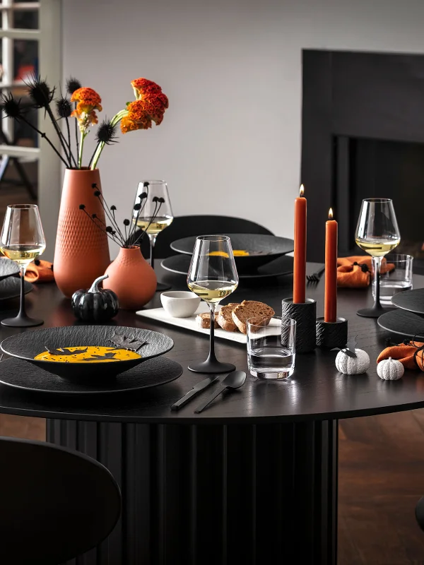 Een moderne eettafel voor vijf personen met zwart Manufacture servies, oranje servetten en herfstdecoraties, waaronder vazen met bloemen en brandende kaarsen, naast een brandende open haard.