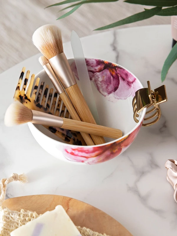 Various make-up brushes arranged in a Rose Garden cereal bowl with floral decoration on a marble tabletop, next to decorative objects.