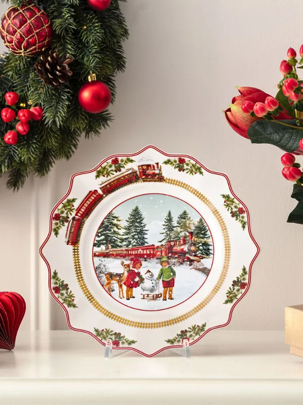 Ein dekorativer Weihnachtsteller mit einer Winterszene wird auf einem Kaminsims mit Weihnachtskränzen, Blumen und eingepackten Geschenken präsentiert.