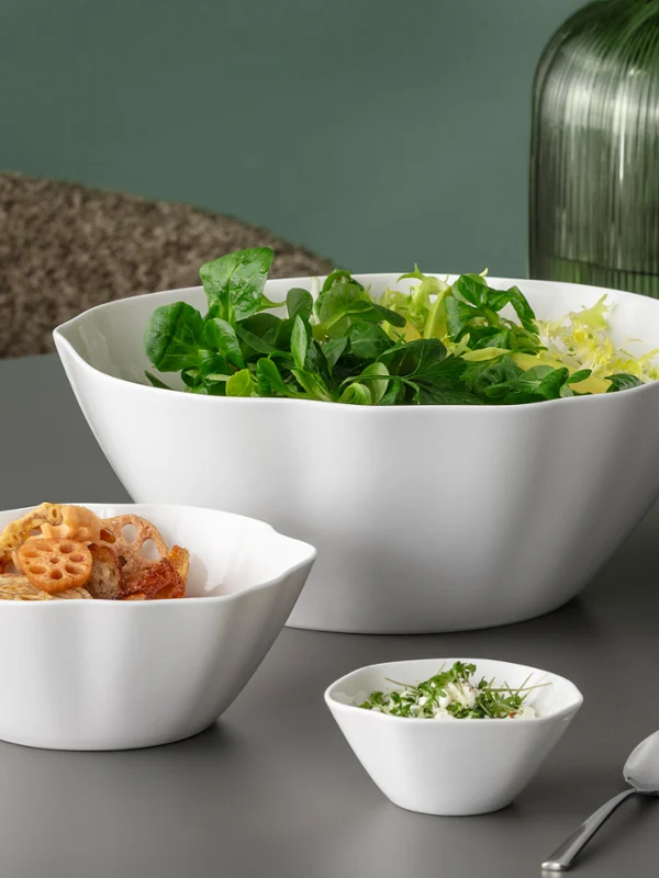 Trois bols blancs Pura contenant de la salade, des pâtes et des herbes hachées sur une table grise, avec des vases verts en arrière-plan.