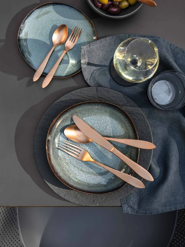 Une décoration de table élégante avec deux assiettes Lave, des couverts de table Manufacture, un plat creux remplis d’olives et un verre de vin blanc sur une nappe grise.