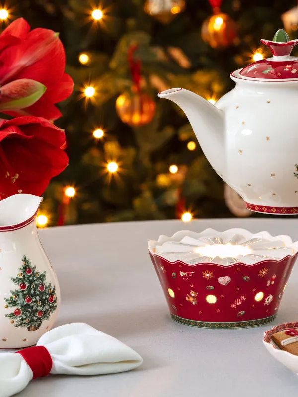 A festive table with a teapot, a warmer, a cup, Christmas ceramics, cookies, and a decorated tree in the background.
