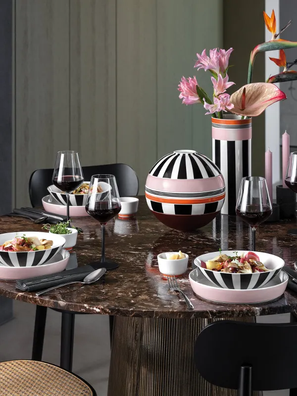 A round marble table set for four, with striped Memphis dinnerware, wine glasses, and a vase with pink flowers.