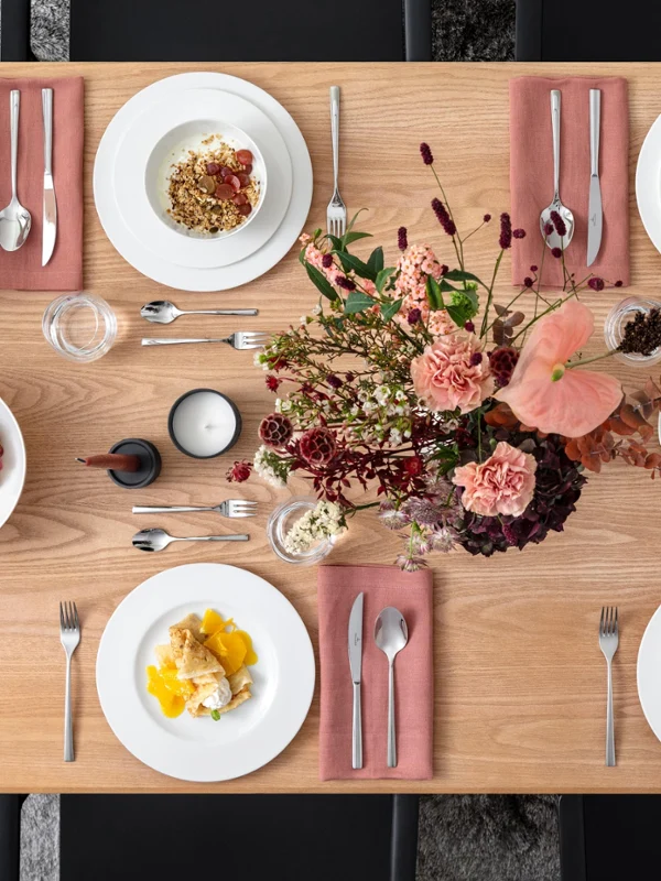 A rectangular wooden table is set for four people. There are plates with food, pink napkins, cutlery, candles, and a floral arrangement on the table. Grapes and spices are also present.