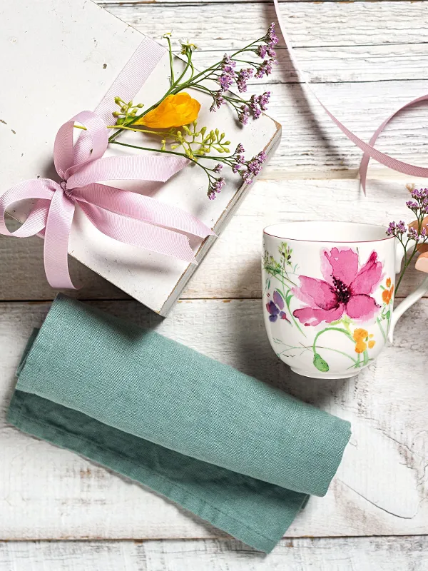 Eine Hand greift nach einem Villeroy & Boch Mariefleur-Becher neben einer Geschenkbox mit einer rosa Schleife und Blumen, die auf einer Holzoberfläche mit einem blaugrünen Tuch steht.