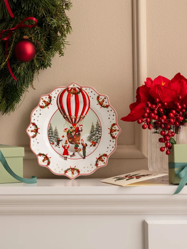 Annual Chistmas edition 2026 plate Ein Weihnachtskranz, ein Dekoteller mit einem Heißluftballon von Villeroy & Boch, grüne Geschenkboxen und rote Blumen, arrangiert auf einem weißen Kaminsims.