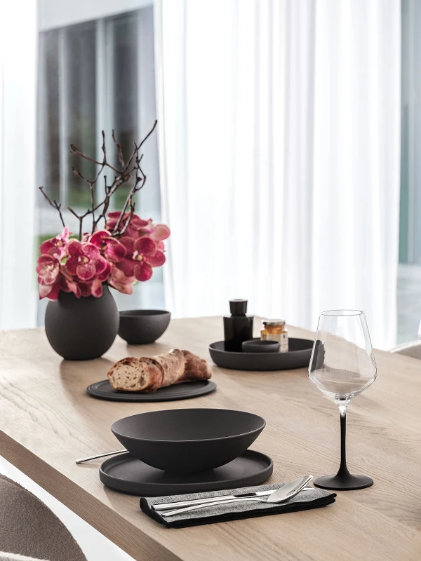 A wooden dining table set with a black La Boule, Manufacture glasses and Piemont flatware from Villeroy & Boch.