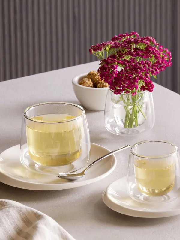 Eine kleine Teekanne und eine Tasse aus Glas von Villeroy & Boch NewMoon stehen auf einem weißen Teller auf einem Tisch neben einer Glasvase mit lila Blumen und einer kleinen Schüssel mit braunen Zuckerwürfeln.