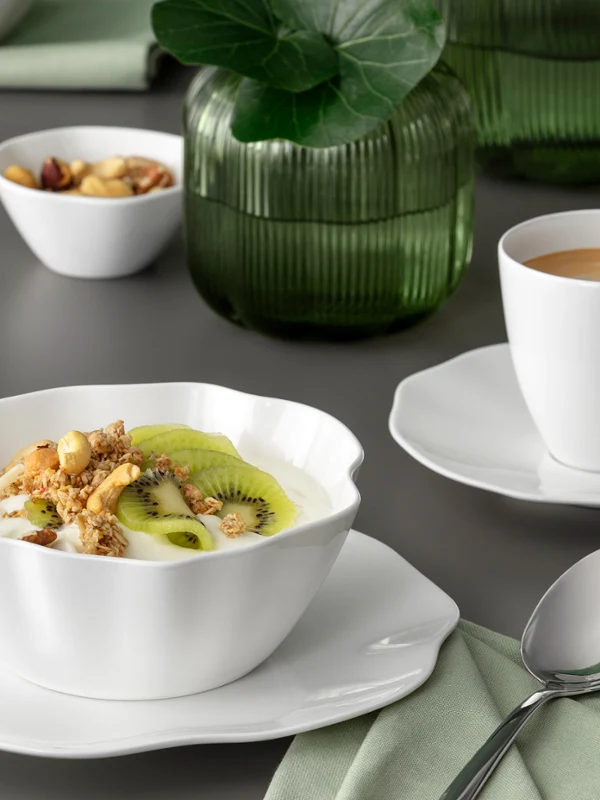 Un bol blanc Pura contenant du muesli et du kiwi, ainsi qu'une tasse de café posée sur une soucoupe, sur une table grise recouverte d'une serviette verte.