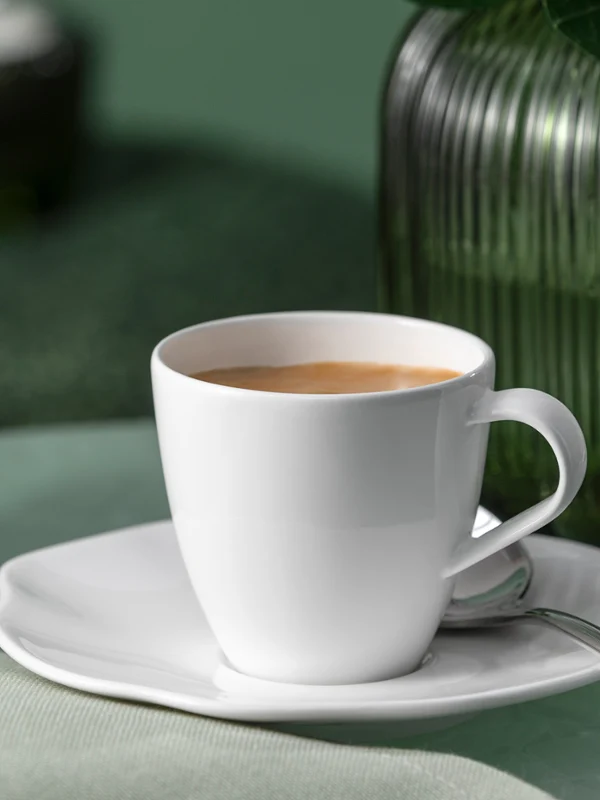 Une tasse à café Pura blanche avec soucoupe et cuillère est posée sur une surface verte à côté d'un vase en verre vert.