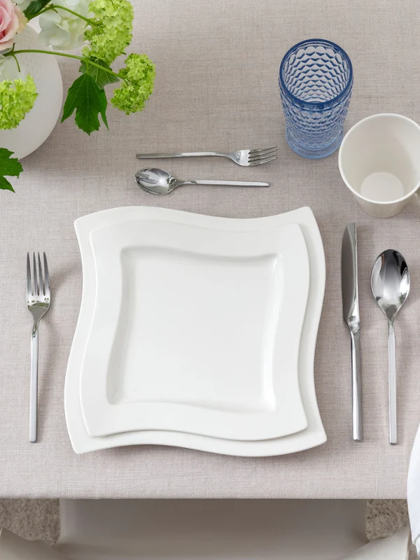 Klare, moderne Draufsicht auf ein einzelnes Gedeck mit NewWave Porzellantellern und Tasse, Boston Blue Longdrinkglas und passendem NewWave Besteck. Neutrale Töne und eine minimalistische Ästhetik vermitteln ein zeitgemäßes Ambiente.