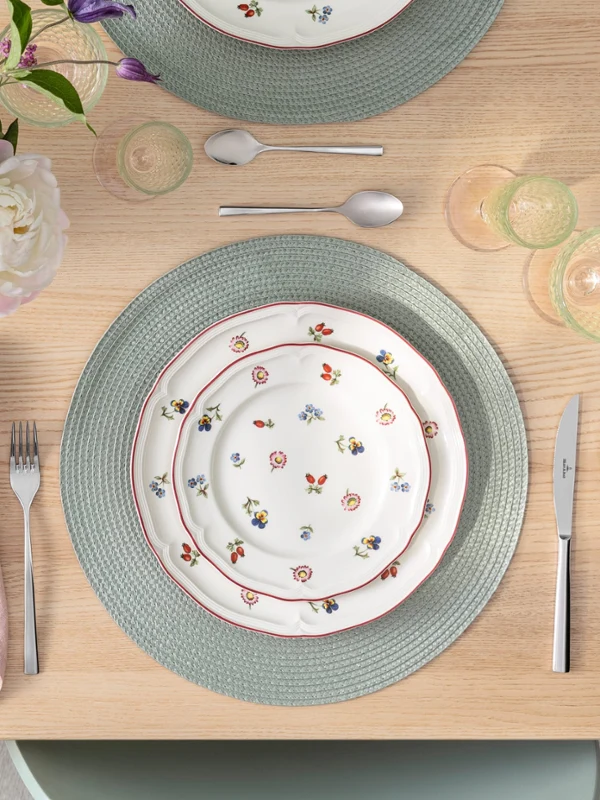 Ein gedeckter Tisch mit Petite Fleur Blumentellern, Besteck, rosa Serviette, grünem Tischset und einem Blumenstrauß auf einem Holztisch.