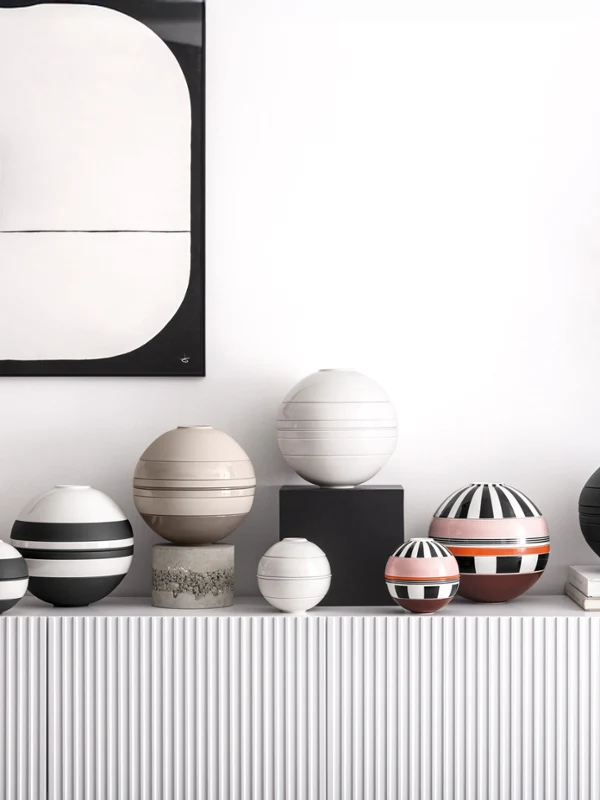 Eine Reihe bunter, gestreifter und gemusterter kugelförmiger La Boule steht auf einer weißen Konsole unter abstrakter Kunst und einer Vase mit orangefarbenen Blumen.