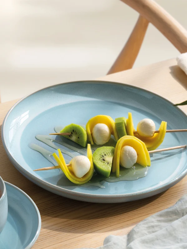 Une assiette bleue perlemor Aqua avec des brochettes de fruits et de mozzarella à côté d'une tasse de café sur une table en bois clair.