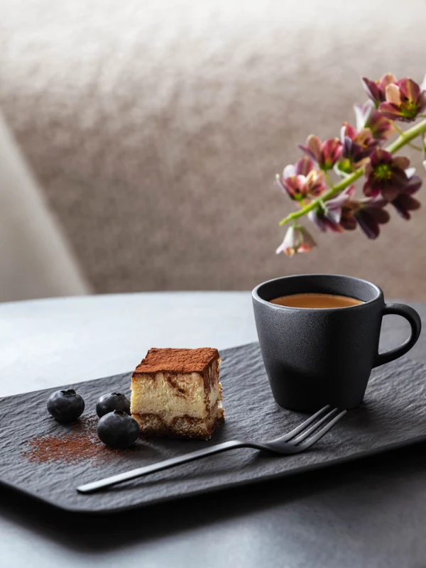 Manufacture-espressokopp og rektangulær tallerken med kake En porsjon tiramisu ved siden av en svart Manufacture-espressokopp, pyntet med blåbær på en moderne, rektangulær Manufacture-tallerken med en dekorativ vase og blomster i bakgrunnen.
