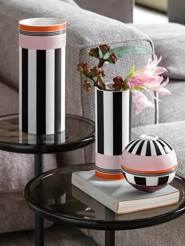 Two black and white striped vases and a round vessel stand on glass tables next to a grey sofa, flowers in a vase