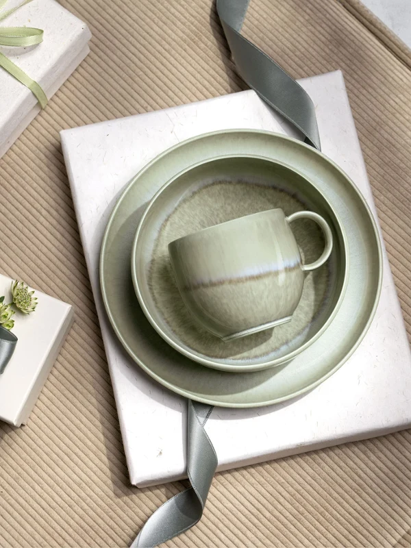 Una tazza, una ciotola e un piatto verdi Perlemor su regali avvolti da nastri grigi, disposti su una superficie beige strutturata.