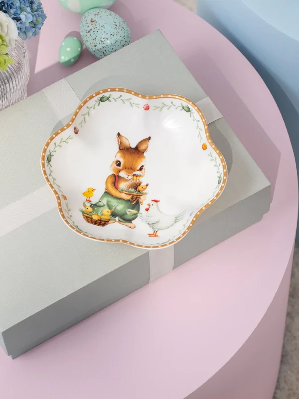 Une boîte cadeau avec un ruban posée sur une table rose, surmontée d’une assiette décorative Annual Easter Edition 2026 avec un lapin et des fleurs.