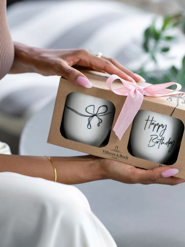 Une personne tient une boîte cadeau avec un ruban rose, à l’intérieur de laquelle se trouvent deux mugs « Happy Birthday ».