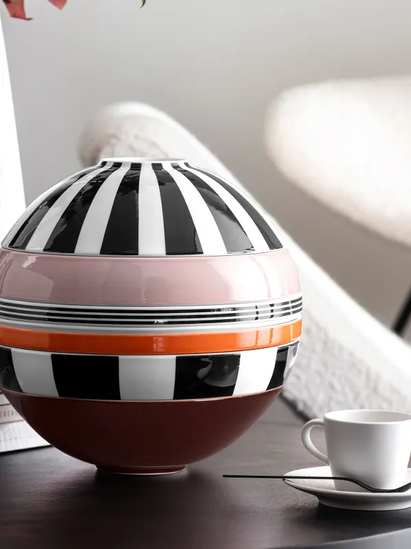 Memphis La Boule with monochrome stripes and a horizontal stripe in pink and orange on a table next to a white cup and saucer, stacked books and a textured white vase. There is a modern chair in the background.