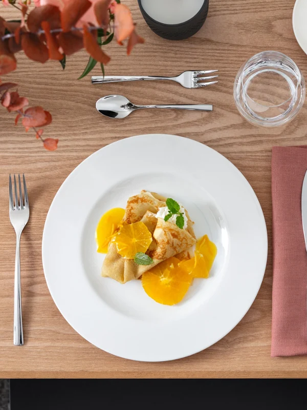 An elegantly set dining table with a plate of crêpes topped with orange slices and mint. Cutlery is placed next to a glass and a bowl with more orange slices.