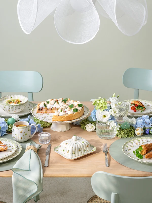 A pastel-themed dining table set for four, featuring plates of food, teacups, blue and white floral arrangements, and elegant tableware, creating a fresh and inviting atmosphere.