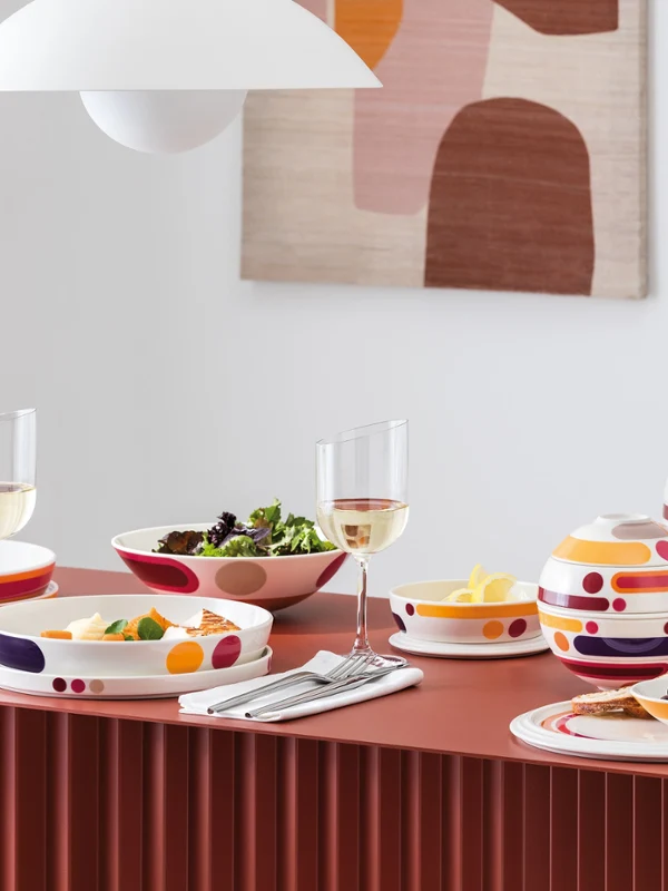 A modern dining table set with colorful patterned dishes, bowls of food, wine glasses, and abstract wall art in the background.