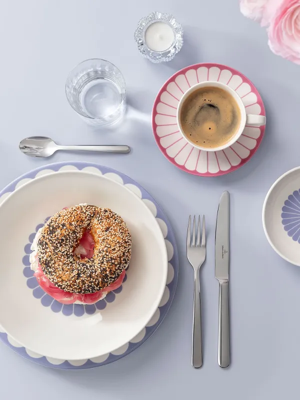 Tischgedeck mit einem Bagel auf einem dekorierten Teller, umgeben von einer Kaffeetasse, kleinen Schüsseln und Besteck auf einer hellblauen Oberfläche mit rosa Blumen an der Seite.