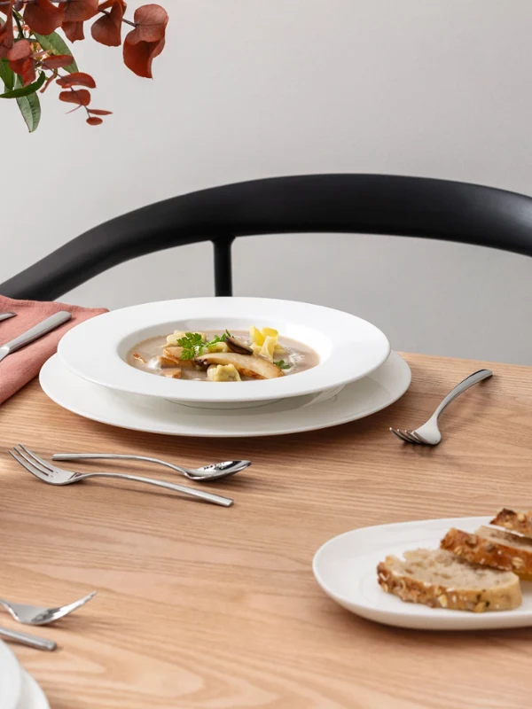 Dining table with a soup bowl and a plate from the Royal collection by Villeroy & Boch. A glass of water and cutlery are arranged on a light-coloured wooden surface. Dark-coloured chair and plant decoration in the background.	