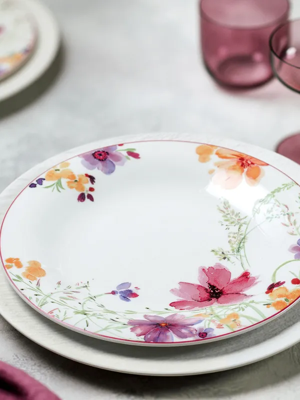 A Mariefleur Basic dinner plate from Villeroy & Boch stands on a white place setting and is accompanied by a purple napkin and a purple glass on the table.