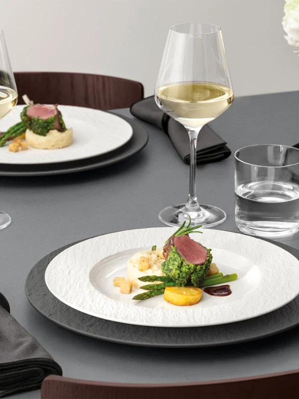A formal table setting with a white plate on a black plate with food on it, glassware, and silver cutlery on a grey table.