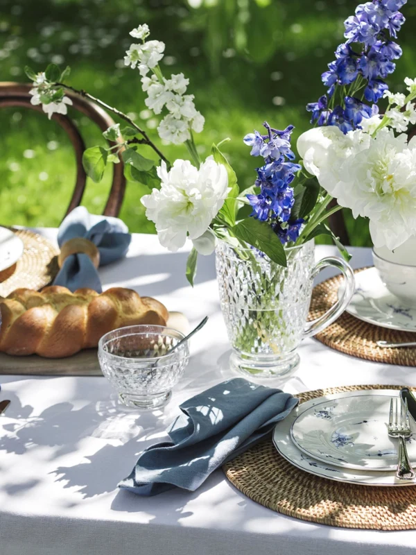 Ein Gartentisch ist mit Blumenporzellan „Alt Luxemburg“ von Villeroy & Boch, Tischsets aus Korbgeflecht, blau getöntem Glasgeschirr, einer Kristallkanne mit blauen und weißen Blumen und einem Brotlaib gedeckt.
