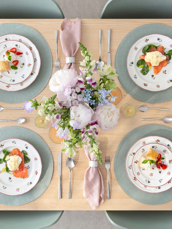 Ein gedeckter Esstisch für vier Personen mit Petite Fleur Blumentellern, rosa Servietten, Getränken und einem Blumenschmuck.