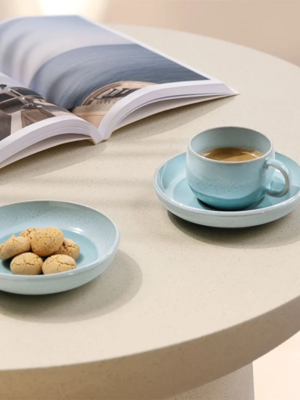 Une tasse à café en nacre, une assiette avec des biscuits et un magazine ouvert sont posés sur une table ronde claire.