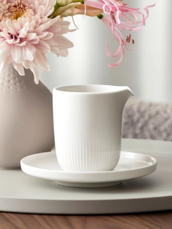 Afina milk jug staged A white ceramic pitcher on a saucer stands on a round tray next to a vase with pink flowers on a wooden table.