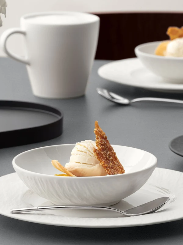 A formal table setting with a Manufacture bowl on top of a plate A formal table setting with a white bowl on a white plate with food on it, cup, and silver cutlery on a grey table.