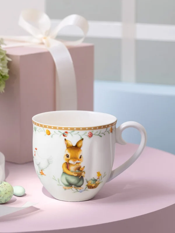 Une tasse décorée Annual Easter Edition posée sur une surface rose à côté d’un ruban blanc, de boîtes-cadeaux pastel et d’un vase de fleurs.