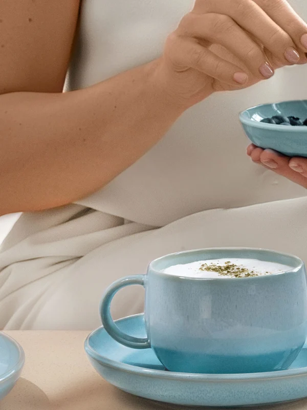 Personne tenant des myrtilles au-dessus d'un bol bleu nacré Aqua, avec une tarte aux myrtilles et une tasse de café nacrée Aqua sur une table devant elle.