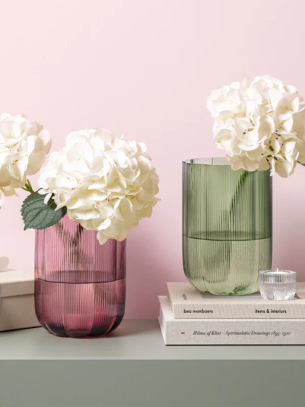 Dos jarrones de cristal Fleur con hortensias blancas sobre una mesa junto a una pila de libros, una caja de regalo y una pequeña lámpara verde.
