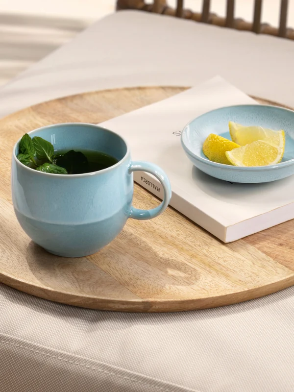 Une tasse bleue nacrée Aqua avec du thé et une soucoupe bleue nacrée Aqua avec des tranches de citron sur un plateau, à côté d'un livre et d'un sac de citrons.