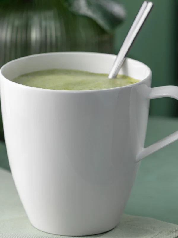 Une tasse blanche Pura Henkelbecher remplie d'une boisson verte et d'une cuillère posée sur une surface claire.