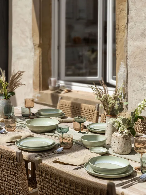 Een zomerse eettafel met groene borden uit de Perlemor-collectie, glazen en bestek op een houten blad. De tafel wordt omlijst door rieten stoelen en het middenstuk is versierd met decoratieve vazen vol bloemen.