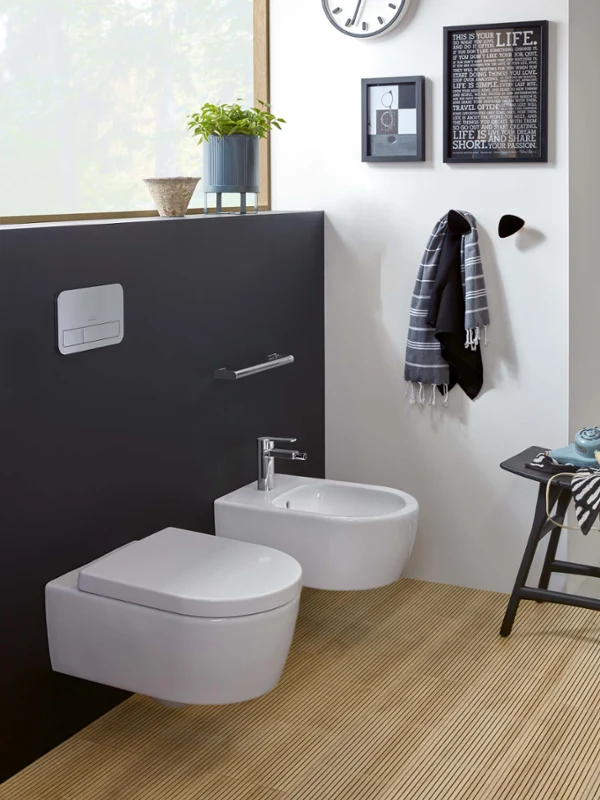 Une salle de bains moderne avec des toilettes et un bidet suspendus de la collection Avento, une serviette sur un crochet, une petite plante sur une étagère et une horloge. Une fenêtre permet à la lumière naturelle de pénétrer.