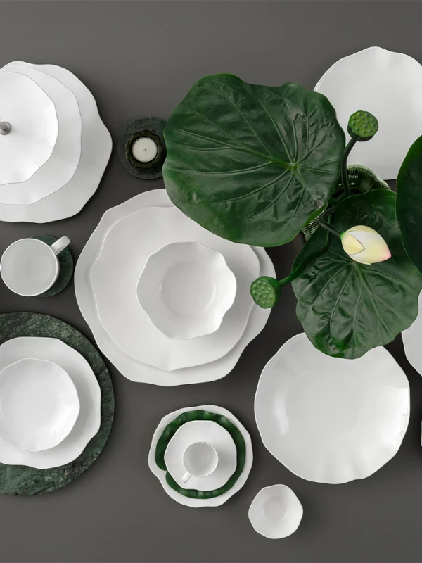 Vaisselle blanche de la collection Pura sur un fond sombre décoré de grandes feuilles vertes et d'une seule fleur blanche.