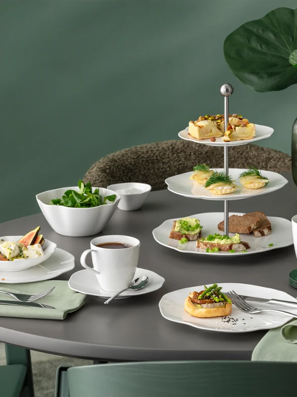 Une table ronde avec de la vaisselle blanche de la collection Pura, divers produits de boulangerie, de la salade, du thé et un décor de feuilles vertes sur fond vert.